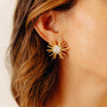 A close-up of a woman's ear wearing a gold starburst stud earring with diamonds, against a soft beige background.