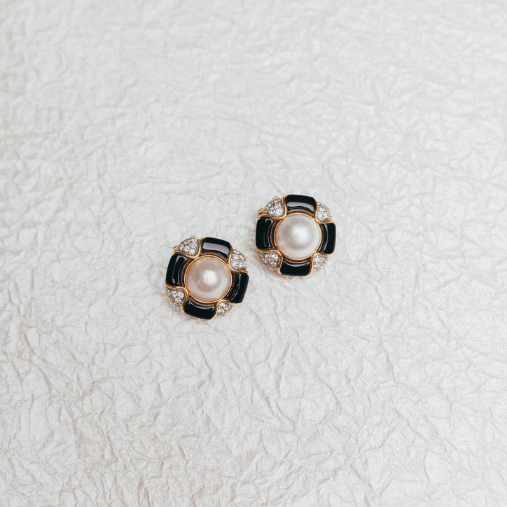 A pair of stud earrings featuring white pearls, black enamel petals, and small diamonds in a gold-tone setting, displayed on a textured off-white surface.