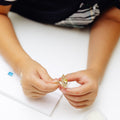 A close-up shows a person in a dark navy shirt holding an ornate gold ring over a white work surface. The ring has a narrow gold band and a large floral or starburst-shaped setting, with a central round yellow-gold stone surrounded by smaller clear, reflective stones arranged like petals. 