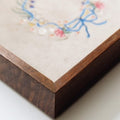 Close-up of a jewelry tray corner with a dark brown wood-grain base and a light beige top featuring embroidered floral and bow details in blue, pink, and white.
