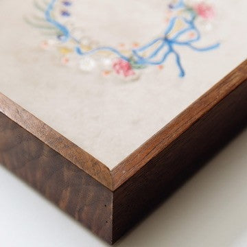 Close-up of a jewelry tray corner with a dark brown wood-grain base and a light beige top featuring embroidered floral and bow details in blue, pink, and white.