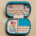 An open turquoise blue zippered jewelry box with cream fabric lining, shown on a textured beige background. The jewelry box contains various colorful jewelry pieces and is decorated with small embroidered motifs.