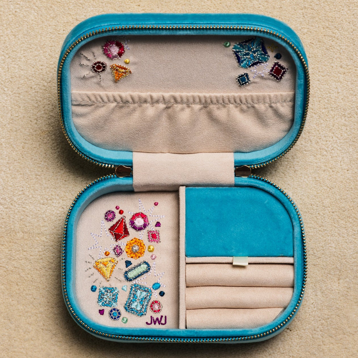 An open turquoise blue zippered jewelry box with cream fabric lining, shown on a textured beige background. The jewelry box contains various colorful jewelry pieces and is decorated with small embroidered motifs.