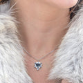 A close-up lifestyle shot of a person wearing a silver heart-shaped pendant necklace with a dark blue heart in the center, styled with a light gray fur coat and gold rings on their fingers.