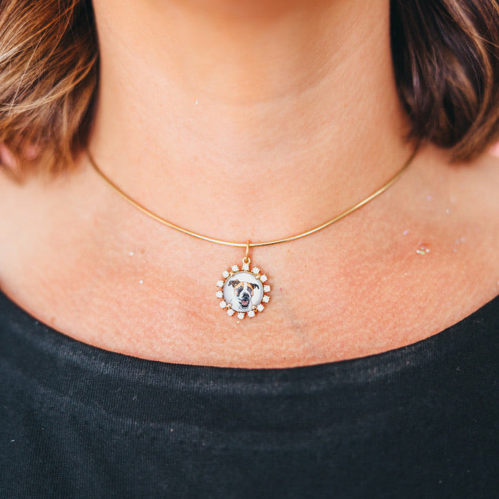 A close-up of a person's neck and chest wearing a delicate gold chain necklace with a custom circular pendant featuring a dog portrait surrounded by small clear stones.