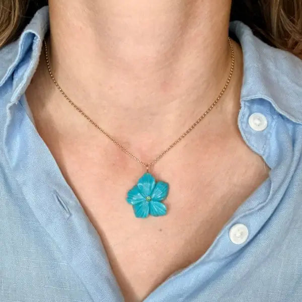 Necklace with a turquoise flower pendant worn by a person in a light blue shirt. Arizona Turquoise Carved Flower. Hand-carved from vibrant Arizona turquoise, this floral pendant is accented with a rich emerald center for a striking pop of color. Finished with a classic 14k gold rolo chain, each piece is naturally unique and designed to be worn every day or layered with ease. Jewelry Dallas.