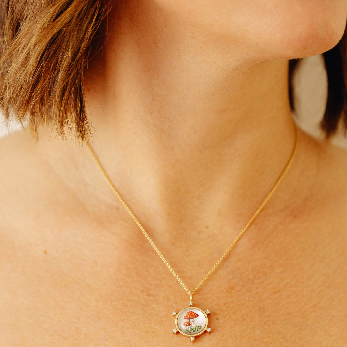 A close-up of a person's neck and chest wearing a delicate gold chain necklace with a circular pendant featuring a small illustrated mushroom design.