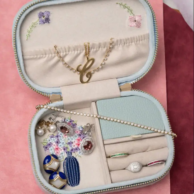 Open blue velvet travel jewelry case with necklaces, rings, and earrings on a pink background. Blooming with Cheer Light Blue Velvet Embroidered Travel Jewelry Box. Jewelry Dallas.