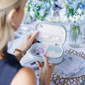 Person holding a jewelry box with floral design, surrounded by flowers and a table setting. Blooming with Cheer Light Blue Velvet Embroidered Travel Jewelry Box. Jewelry Dallas.