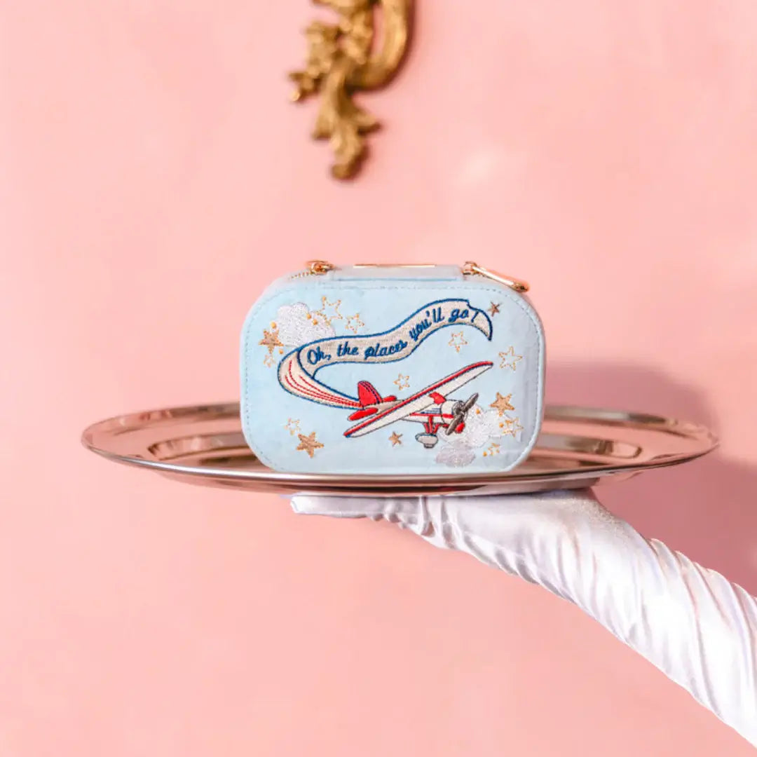 Light blue velvet travel jewelry box with airplane design on a pink background. Bon Voyage Light Blue Velvet Embroidered Travel Jewelry Box. Jewelry Dallas.