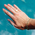 A close-up of a person's hand resting in turquoise water, wearing a gold ring with a white rectangular stone and a gold chain bracelet.