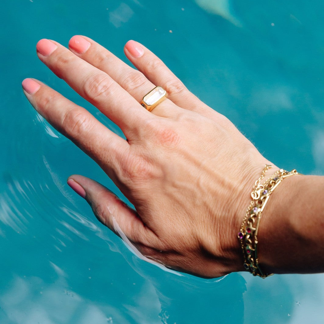 A close-up of a person's hand resting in turquoise water, wearing a gold ring with a white rectangular stone and a gold chain bracelet.