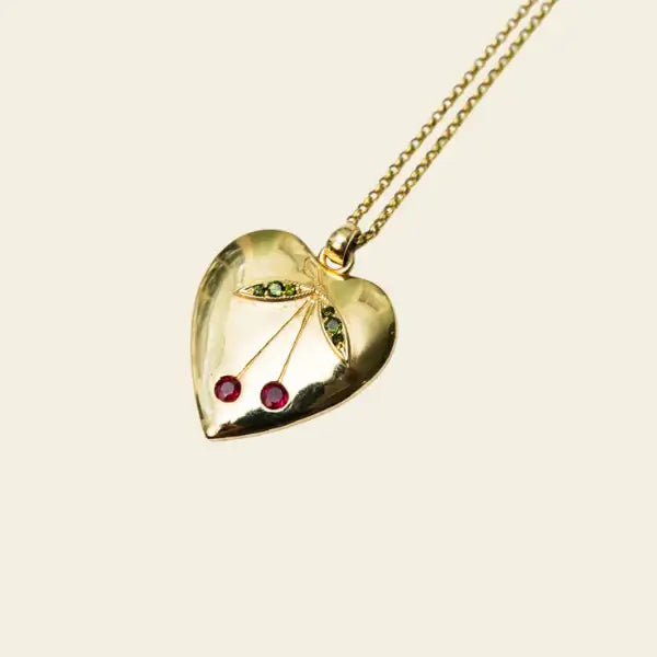 Gold heart-shaped pendant with natural gemstones in the shape of a cherry on a beige background. Cherry Heart Pendant. 14K gold heart pendant features a charming cherry motif brought to life with vibrant gemstones. Jewelry Dallas.