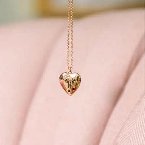 Gold heart-shaped pendant with natural gemstones in the shape of a cherry on a beige background. Cherry Heart Pendant. 14K gold heart pendant features a charming cherry motif brought to life with vibrant gemstones. Jewelry Dallas.