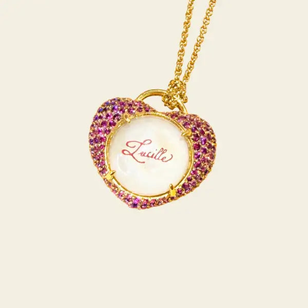 Gold necklace with a heart-shaped pendant featuring the name 'Lucille' on a beige background. Custom Calligraphied Essex Crystal. Materials: Solid 14kt Gold Frame with Hand-Carved Rock Crystal and Natural Pink Sapphires
Customization: Single Initial or Monogram, Reverse-Carved and Hand-Painted in Custom Calligraphy
Optional: Diamond Accents Available Upon Request
Chain: Sold Separately (Pairs Beautifully with Our Classic Cable or Paperclip Chains)
Origin: Handcrafted in the USA. Jewelry Dallas.