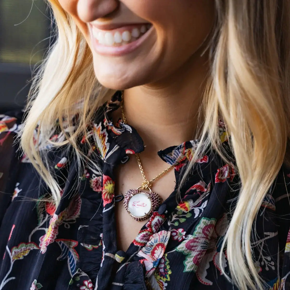 A woman wearing a floral blouse and a necklace with a calligraphed pendant. Custom Calligraphied Essex Crystal. Materials: Solid 14kt Gold Frame with Hand-Carved Rock Crystal and Natural Pink Sapphires
Customization: Single Initial or Monogram, Reverse-Carved and Hand-Painted in Custom Calligraphy
Optional: Diamond Accents Available Upon Request
Chain: Sold Separately (Pairs Beautifully with Our Classic Cable or Paperclip Chains)
Origin: Handcrafted in the USA. Jewelry Dallas.