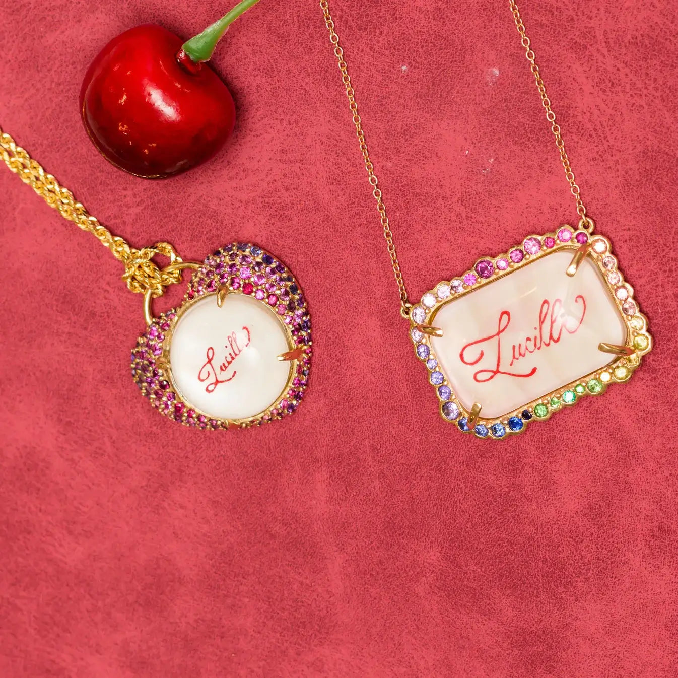 Two necklaces with 'Lucille' calligraphed on them, surrounded by natural colored gemstones. Custom Calligraphied Essex Crystal. Materials: Solid 14kt Gold Frame with Hand-Carved Rock Crystal and Natural Pink Sapphires
Customization: Single Initial or Monogram, Reverse-Carved and Hand-Painted in Custom Calligraphy
Optional: Diamond Accents Available Upon Request
Chain: Sold Separately (Pairs Beautifully with Our Classic Cable or Paperclip Chains)
Origin: Handcrafted in the USA. Jewelry Dallas.