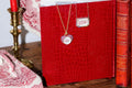 Red embossed book with a necklace on top, next to a candle and another book. Custom Calligraphied Essex Crystal. Materials: Solid 14kt Gold Frame with Hand-Carved Rock Crystal and Natural Pink Sapphires
Customization: Single Initial or Monogram, Reverse-Carved and Hand-Painted in Custom Calligraphy
Optional: Diamond Accents Available Upon Request
Chain: Sold Separately (Pairs Beautifully with Our Classic Cable or Paperclip Chains)
Origin: Handcrafted in the USA. Jewelry Dallas.