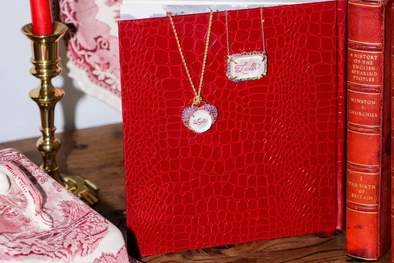Red embossed book with a necklace on top, next to a candle and another book. Custom Calligraphied Essex Crystal. Materials: Solid 14kt Gold Frame with Hand-Carved Rock Crystal and Natural Pink Sapphires
Customization: Single Initial or Monogram, Reverse-Carved and Hand-Painted in Custom Calligraphy
Optional: Diamond Accents Available Upon Request
Chain: Sold Separately (Pairs Beautifully with Our Classic Cable or Paperclip Chains)
Origin: Handcrafted in the USA. Jewelry Dallas.