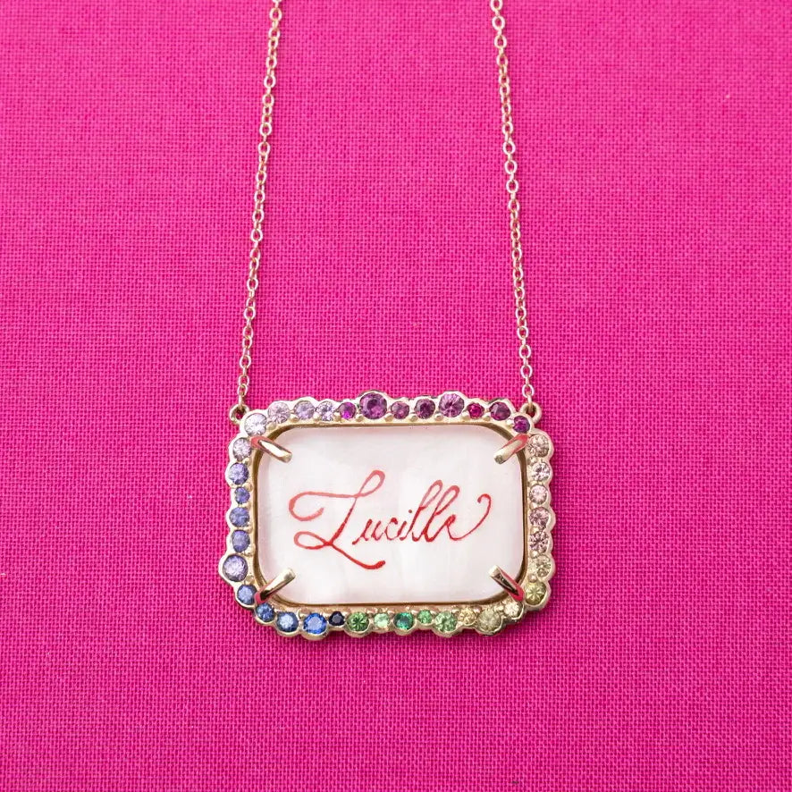 Necklace with a pendant displaying the name 'Lucille' on a pink background. Custom Calligraphied Essex Crystal. Materials: Solid 14kt Gold Frame with Hand-Carved Rock Crystal and Natural Pink Sapphires
Customization: Single Initial or Monogram, Reverse-Carved and Hand-Painted in Custom Calligraphy
Optional: Diamond Accents Available Upon Request
Chain: Sold Separately (Pairs Beautifully with Our Classic Cable or Paperclip Chains)
Origin: Handcrafted in the USA. Jewelry Dallas.