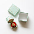 A mint green textured jewelry box set on a white background, featuring one closed box, one open box with a red velvet interior and a gold starburst brooch, and one empty box.