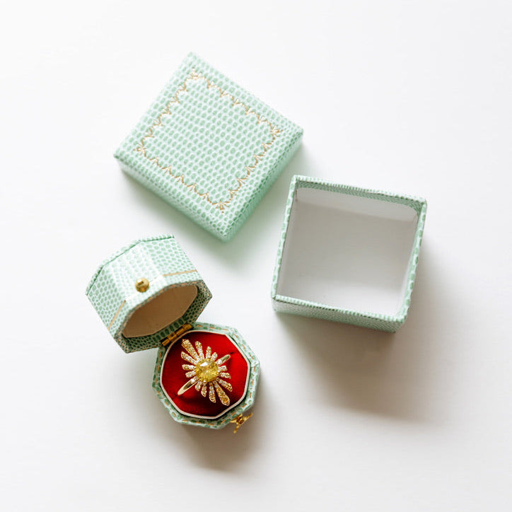 A mint green textured jewelry box set on a white background, featuring one closed box, one open box with a red velvet interior and a gold starburst brooch, and one empty box.
