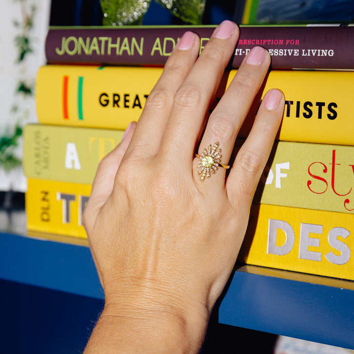 A yellow gold sunburst ring featuring a large round yellow center stone and smaller yellow and clear accent stones, set against a solid cream background.
