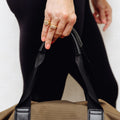 A close-up of a person's hand holding the handle of a brown canvas gym bag with black straps. The person is wearing black leggings and gold rings.