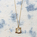A gold pendant necklace featuring a crescent moon and stars design, displayed on a textured, off-white paper background with blue watercolor-like splashes.