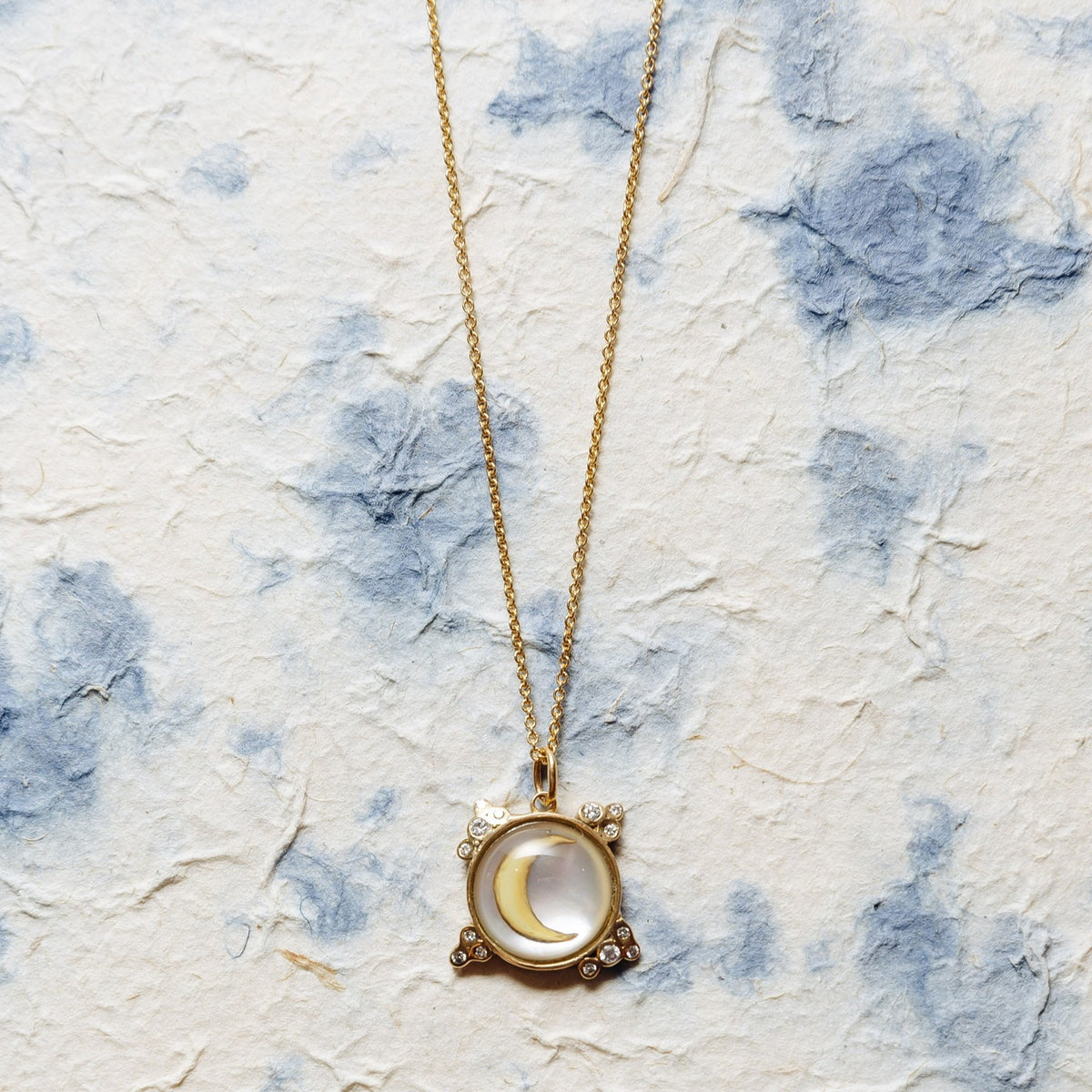 A gold pendant necklace featuring a crescent moon and stars design, displayed on a textured, off-white paper background with blue watercolor-like splashes.