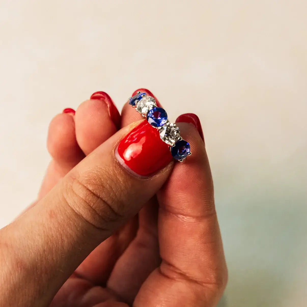 Hand holding ring with sapphires and diamonds on a beige background. Five Stone Blue Sapphire and Diamond Ring. # of Diamonds:2 Carat Weight:0.60 carat Shape:RoundColor:G-HClarity:SI1-SI2Cut:Excellent # of Sapphires:3Carat Weight:2.10 carats Shape:RoundColor:BlueClarity:Eye CleanCut:Excellent. 14k gold. Jewelry Dallas.