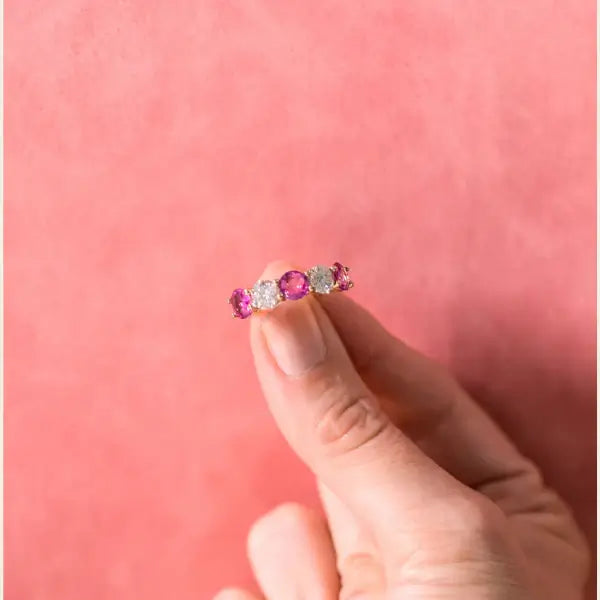 Gold ring with pink sapphires and diamonds held between fingers against a pink background. Five Stone Pink Sapphire and Diamond Ring. # of Diamonds:2.5 Carat Weight:1.60 carats Shape:RoundColor:G-HClarity:SI1-SI2Cut:Excellent # of Sapphires:3Carat Weight:2.40 carats Shape:RoundColor:PinkClarity:Eye CleanCut:Excellent. Jewelry Dallas.