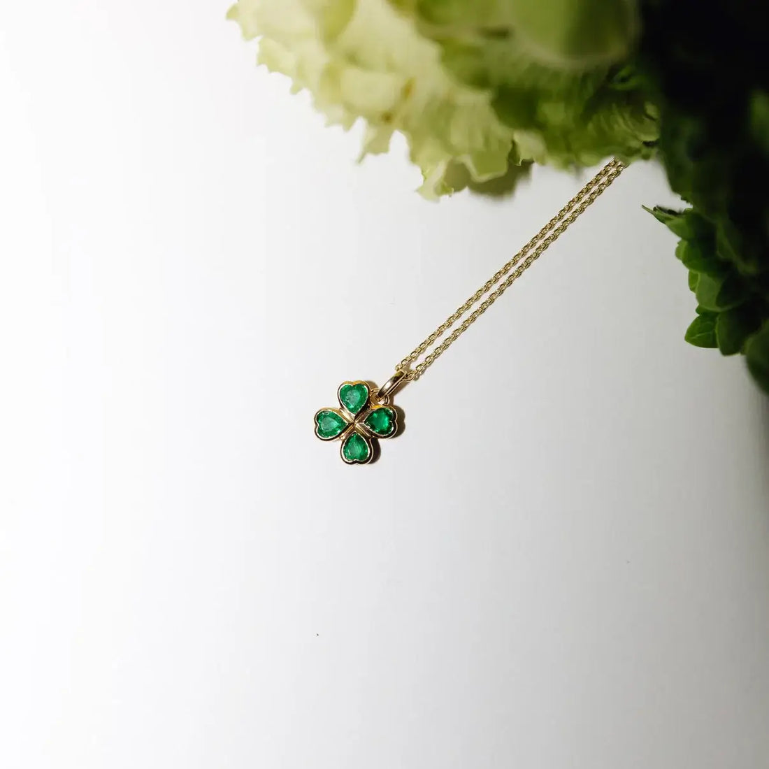 Gold necklace with a green four-leaf clover pendant on a beige background. Four Heart Clover. Crafted in 14k gold and set with 0.74 carats of sparkling natural emeralds. Jewelry Dallas.