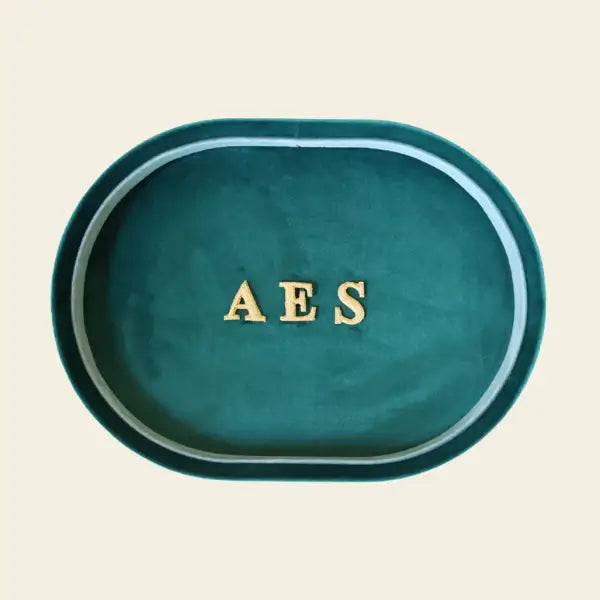 Green oval velvet jewelry tray with 'AES' letters on a beige background. Green Velvet Jewelry Tray. Jewelry Dallas.