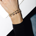 A close-up of a person's wrist wearing two delicate gold bracelets adorned with small heart charms and colorful stones, paired with a black ribbed long-sleeve top against a white and black background.