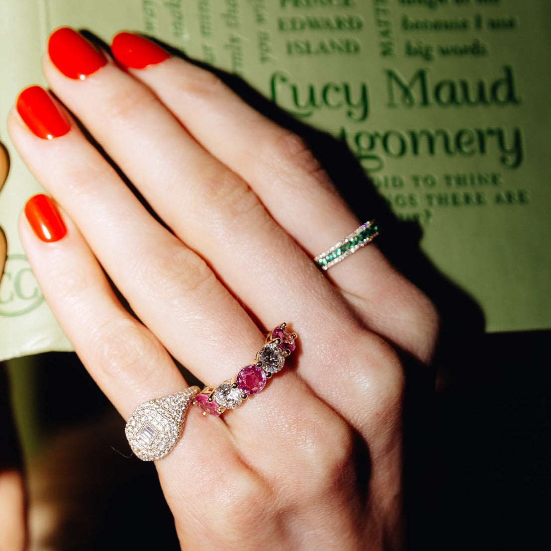 Close-up of a person's hands with bright red nail polish, wearing multiple rings including a gold ring with pink sapphires and diamonds, a silver-toned ring with a clear stone, and a green band, holding a book with a green cover.