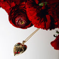 A gold heart-shaped pendant with natural gemstones in the shape of a cherry, hanging from a delicate gold chain next to a bouquet of deep red roses on a white surface.