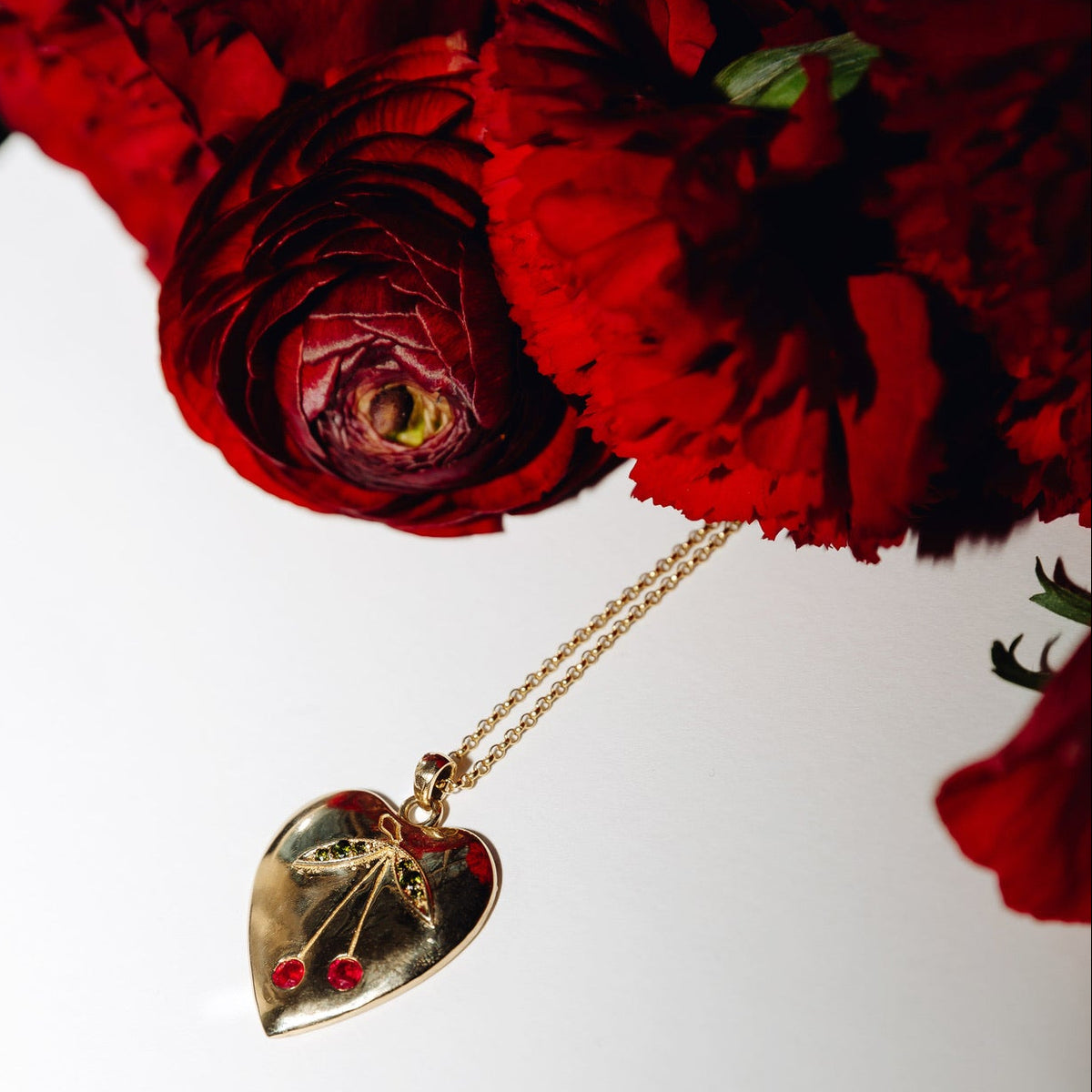 A gold heart-shaped pendant with natural gemstones in the shape of a cherry, hanging from a delicate gold chain next to a bouquet of deep red roses on a white surface.