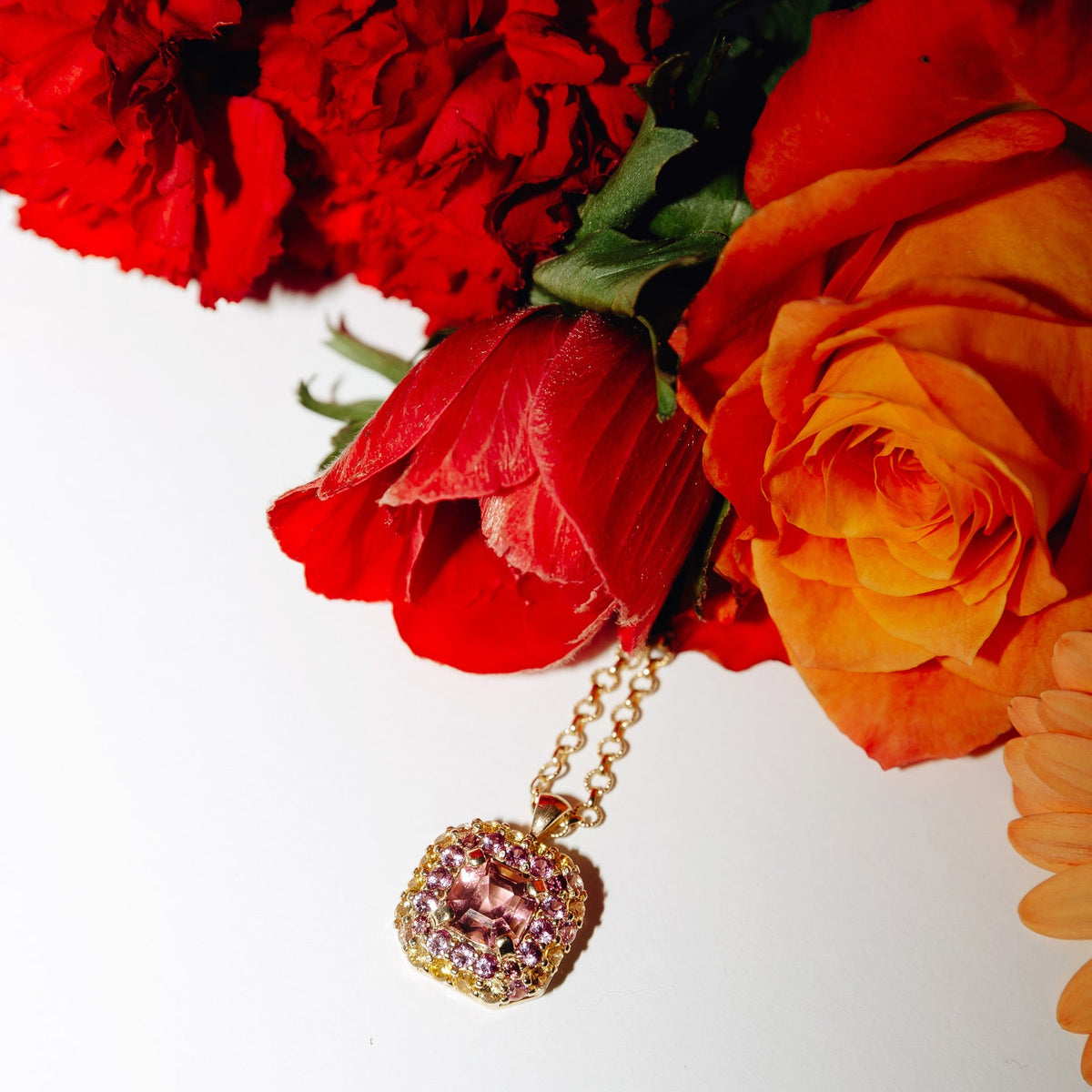 Gold necklace with pink gemstones in front of red and orange flowers on a white background