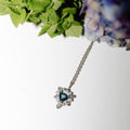 A silver-toned pendant necklace with a heart-shaped blue gemstone surrounded by small clear stones, hanging against a white background with decorative green and blue-purple flowers at the top.