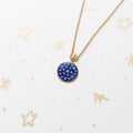 A gold chain necklace with a round blue and gold pendant featuring the star field, displayed on a white background with subtle gold star patterns.