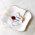 A styled flat-lay of gold and silver jewelry displayed on a white ceramic dish with gold trim and a blue monogram. The jewelry includes a gold necklace with a red circular pendant, a gold statement earring with colorful crystals, and a silver ring with a clear gemstone, all set on a light marble surface.