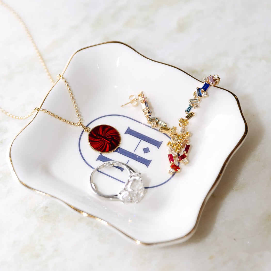 A styled flat-lay of gold and silver jewelry displayed on a white ceramic dish with gold trim and a blue monogram. The jewelry includes a gold necklace with a red circular pendant, a gold statement earring with colorful crystals, and a silver ring with a clear gemstone, all set on a light marble surface.