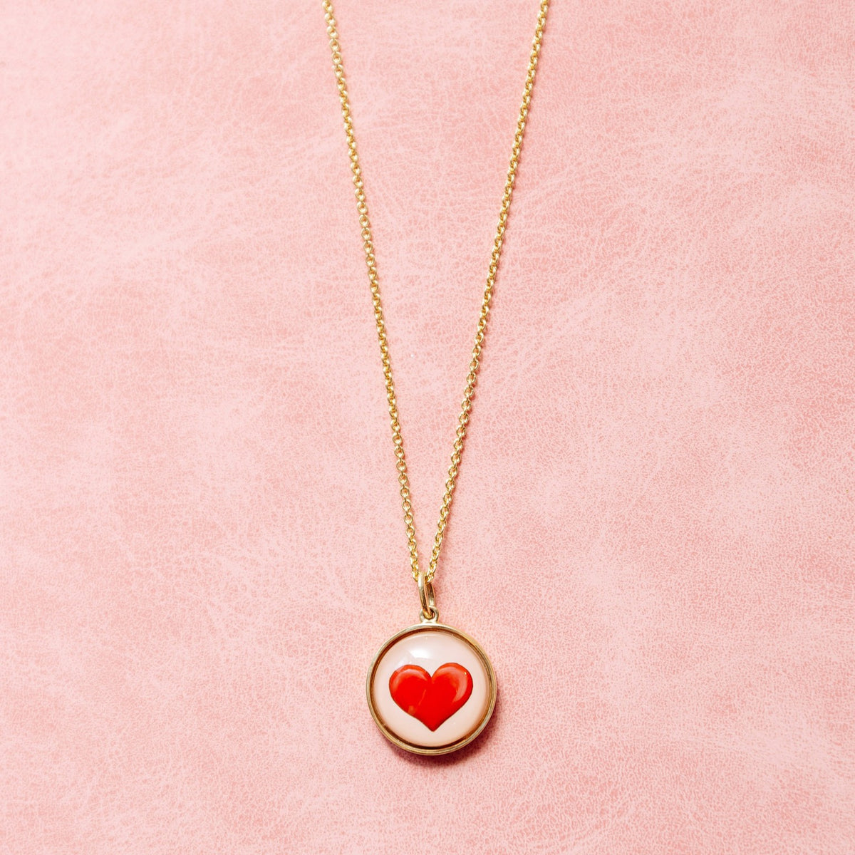 A gold chain necklace with a round pendant featuring a red heart on a white background, displayed against a textured pink backdrop.