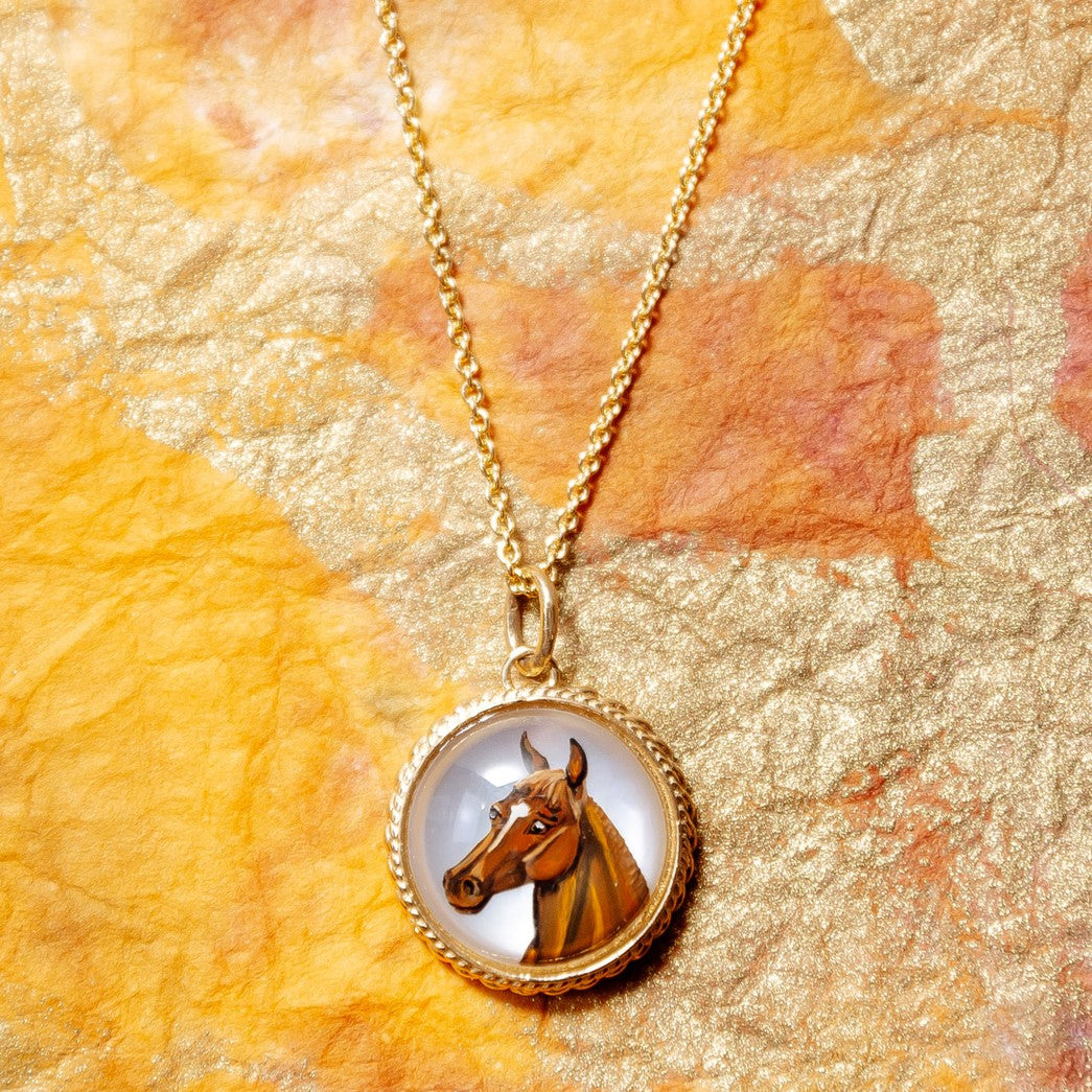 A gold chain necklace with a round pendant featuring a painted brown horse head on a white background, displayed on a textured orange and gold paper surface.