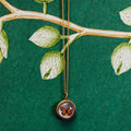 A gold pendant necklace featuring a butterfly preserved inside a circular dome, hanging against a dark green background with a painted leafy branch pattern.