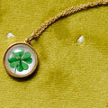 A gold chain necklace with a round pendant featuring a green four-leaf clover under a clear dome, displayed on a textured olive green velvet background.