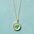 A gold chain necklace with a round pendant featuring a green four-leaf clover under clear cover, displayed on a textured light blue background.