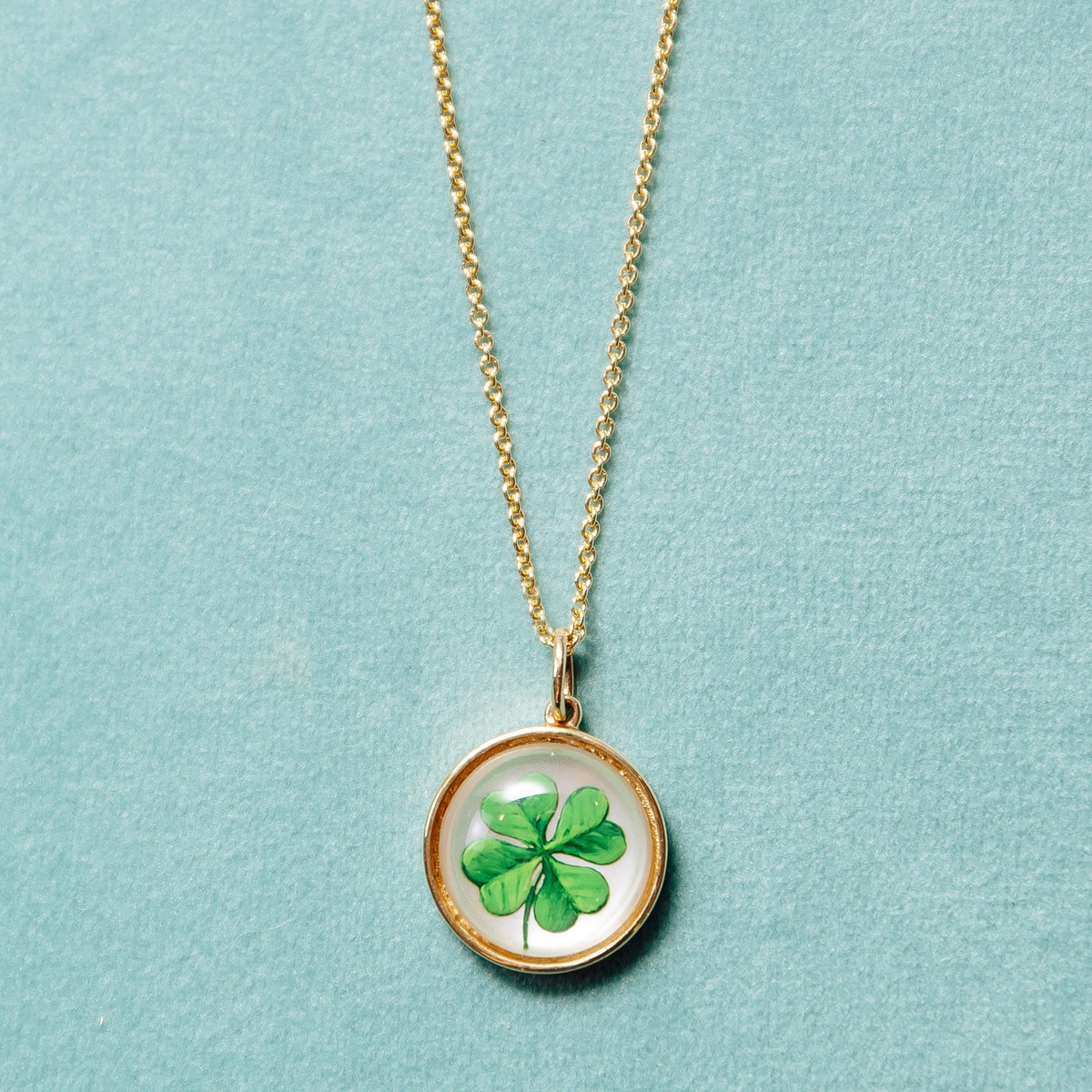 A gold chain necklace with a round pendant featuring a green four-leaf clover under clear cover, displayed on a textured light blue background.