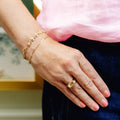 A close-up of a person's hand wearing a wide gold ring with a clear rectangular gemstone, along with two delicate gold bracelets and a pink top.